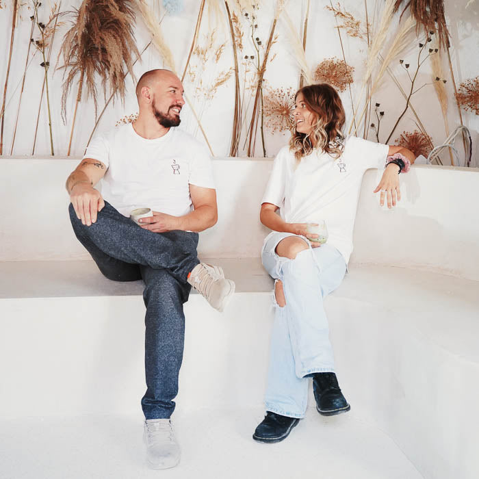 Flo und Sunny vom unbound coffee shop sitzen mit Kaffeetassen vor einer Wand mit Trockenblumen